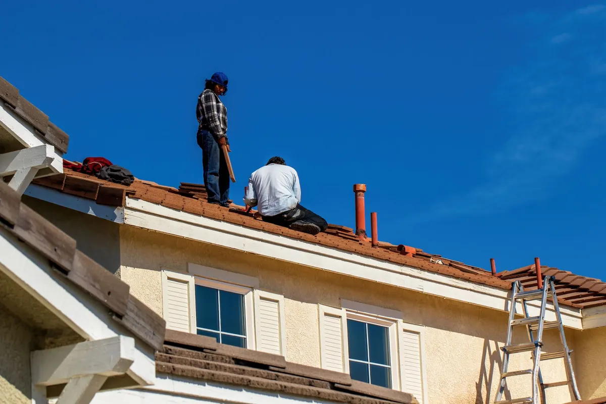 Two roofers working on the tile roof of a stucco home, with a ladder at the eave