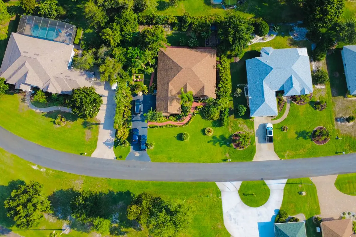 Aerial view of single-story homes with tile and metal roofs on green lots along a curving road